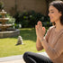 A woman sits cross-legged outdoors, eyes closed, smiling peacefully, wearing an Evil Eye Bracelet as a good luck charm in a serene garden.
